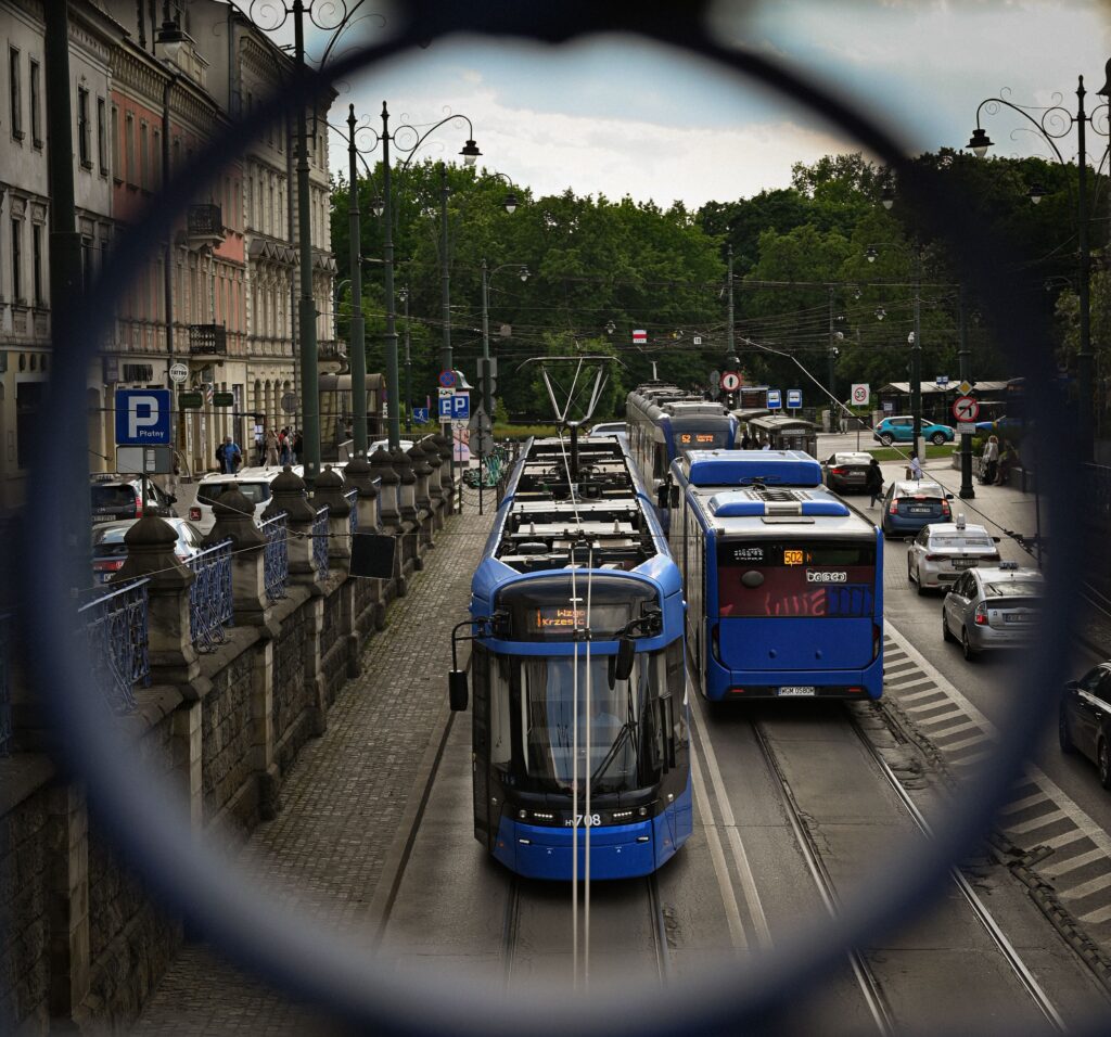 A blue tram and bus navigate a bustling street in Kraków, framed through a circular structure.